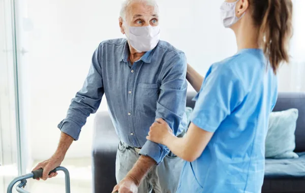 Foto de un señor mayor andando con un andarin acompañado por una enfermera