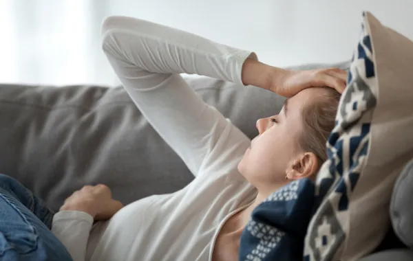 Foto de una mujer joven tumbada en un sofá y con la mano en la cabeza por el dolor