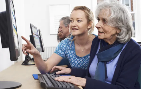 Foto de una mujer mayor en el ordenador acompañada por una chica joven que le explica algo