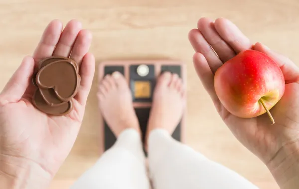 Foto de una persona subida en un báscula que sostiene en una mano una manzana y en otra chocolate