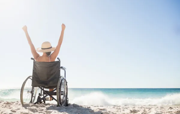 Foto de una chica en silla de ruedas en la playa mirando al mar y con los brazo en alto
