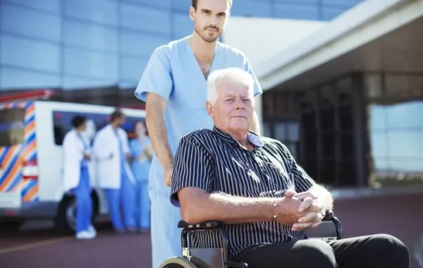 Hombre mayor en silla de ruedas acompañado por un profesional hospitalario en la entrada del Hospital