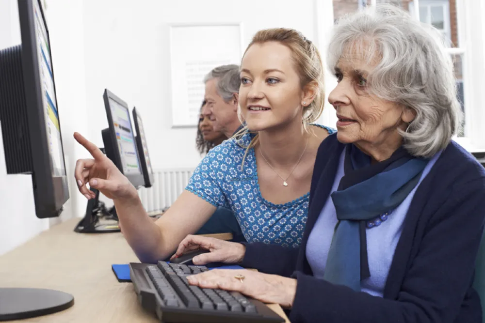 Foto de una mujer mayor en el ordenador acompañada por una chica joven que le explica algo