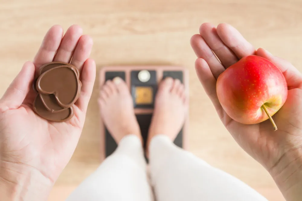 Foto de una persona subida en un báscula que sostiene en una mano una manzana y en otra chocolate
