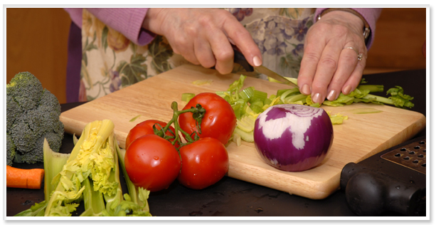 Las manos de una mujer mayor cortan apio en una tabla de cocina, junto a la cual hay tomates, cebolla, brócoli