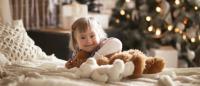 Foto de un niña con sindrome de down sentada en una cama con un arbol de navidad de fondo