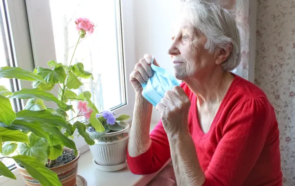 Foto de una señora mayor mirando por la ventana y sosteniendo en una mano una mascarilla