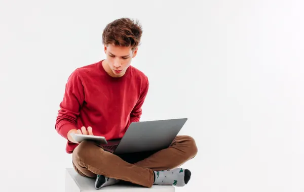 Foto de un chico joven con un ordenador y consultando un movil