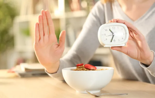 Foto de la mano de una mujer en señal de stop frente a un bol de comida y con la otra mano sujeta un despertador