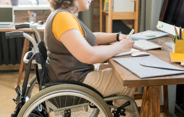 Foto de un chica joven en silla de ruedas sentada en un escritorio y escribiendo
