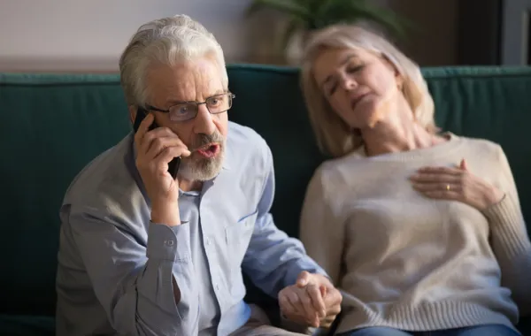 Foto de una pareja adulta en la que la mujer está sentada con los ojos cerrado y el hombre llamando por telefono con cara que expresa urgencia