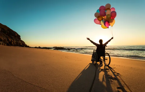 Foto de una chica en silla de ruedas que sostiene unos globos en paisaje de una playa