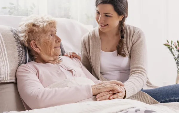 Foto de un señora mayor tumbada en la cama acompañada por una chica joven que le coge de la mano