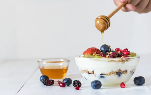 Foto de un mano que vierte miel con un palito en un postre de yougurt y frutas
