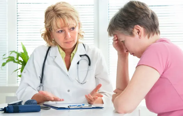 Foto de una mujer con mano en la cabeza con cara de dolor acompañada de una médico que le atiende