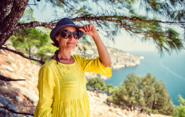 Foto de una mujer adulta con gafas de sol y sombrero debajo de un arbol cerca de una playa