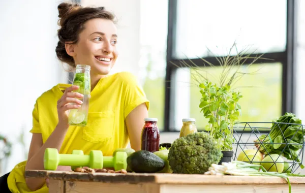 Foto de una mujer joven sonriendo y sentada en una mesa con alimentos sanos y bebiendo agua