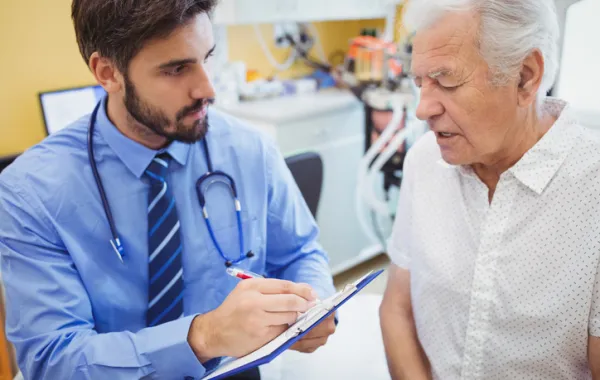 Foto en un hospital de un señor mayor acompañado de uno joven que toma notas en 