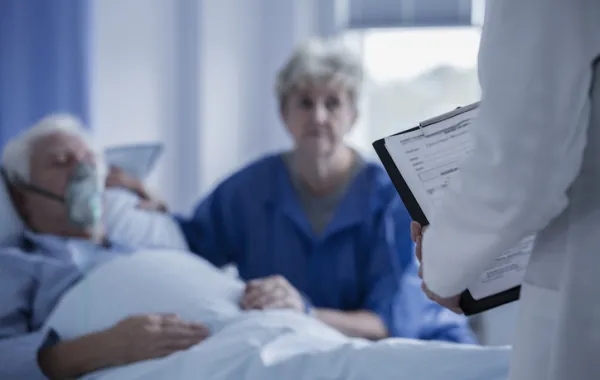 Foto de un hombre tumbado en la cama con oxígeno acompañado de su mujer que escucha a un médico