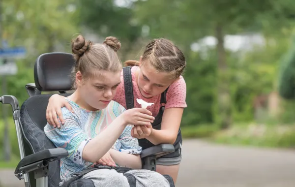 Foto de dos niñas una en silla de ruedas y acompañada de otra niña que le enseña un dibujo