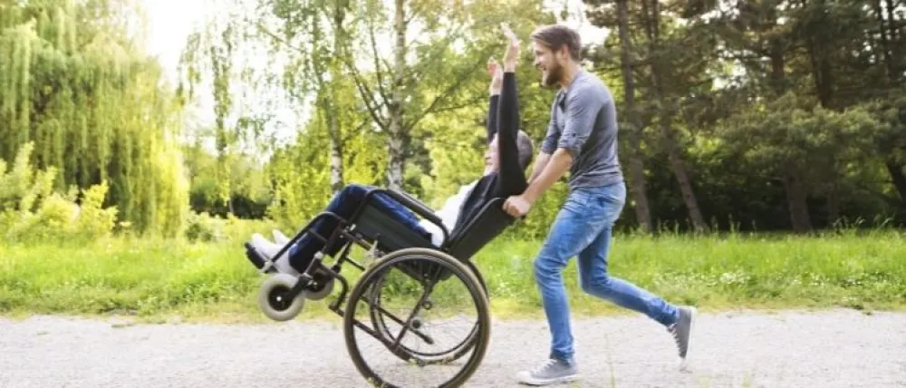 Foto de un señor mayor en silla de ruedas que empuja un chico joven en un parque y con sonrisa de alegria