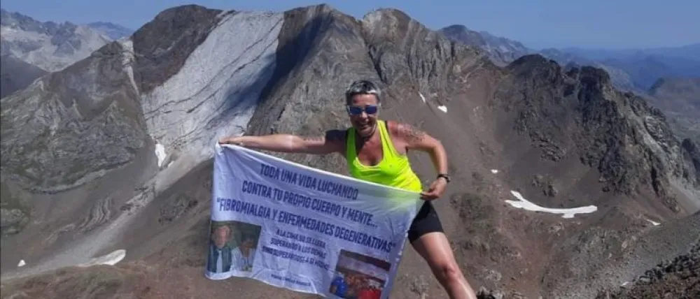Foto de Maria en el pico de una montaña sosteniendo un cartel con la foto de varias personas y frases como fibromialgia y enfermedades degenerativas