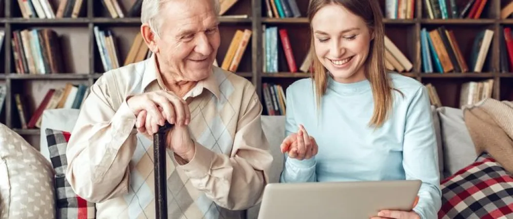 Foto de un señor mayor sonriendo con una chica joven que le explica algo delante de un ordenador
