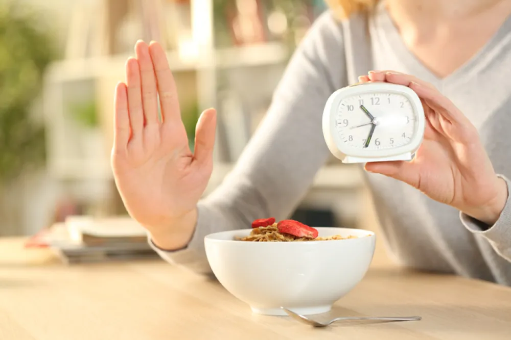 Foto de la mano de una mujer en señal de stop frente a un bol de comida y con la otra mano sujeta un despertador