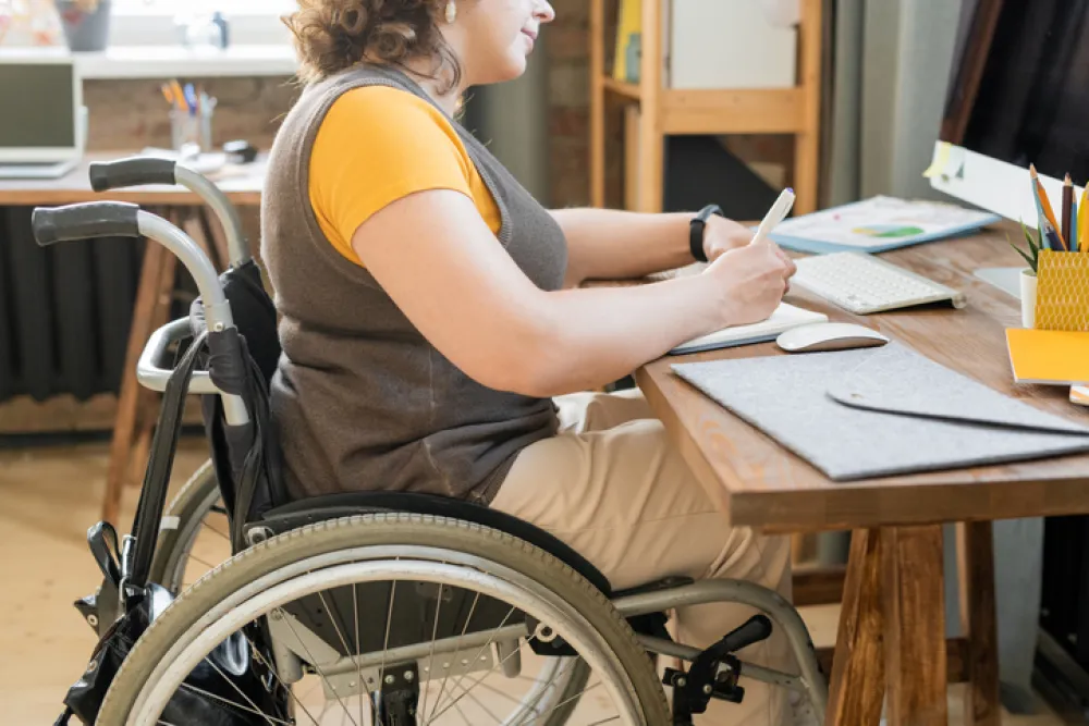 Foto de un chica joven en silla de ruedas sentada en un escritorio y escribiendo