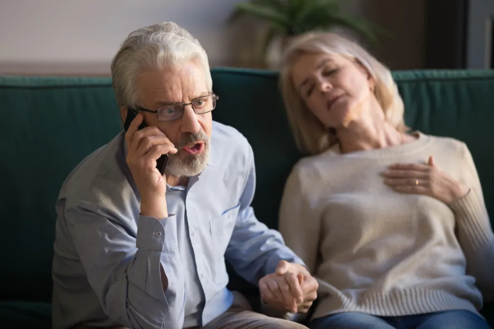 Foto de una pareja adulta en la que la mujer está sentada con los ojos cerrado y el hombre llamando por telefono con cara que expresa urgencia