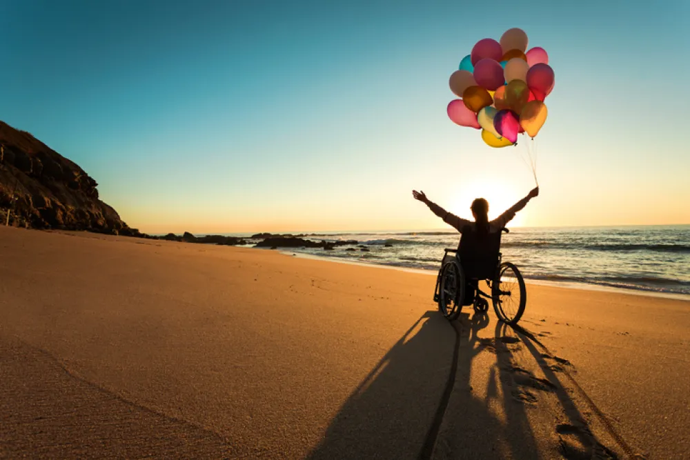 Foto de una chica en silla de ruedas que sostiene unos globos en paisaje de una playa